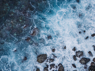 Aerial top-down view of turquoise ocean waves crashing against a rugged rocky shoreline. The dynamic movement of white sea foam contrasts with dark volcanic stones, creating a dramatic natural texture © luciano