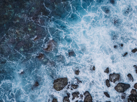 Aerial top-down view of turquoise ocean waves crashing against a rugged rocky shoreline. The dynamic movement of white sea foam contrasts with dark volcanic stones, creating a dramatic natural texture