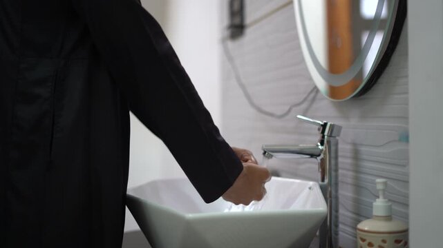 4K Close up of Muslim man in black jubbah lifting faucet lever to start water flow and washing hands in modern white basin, hygiene and lifestyle concept.