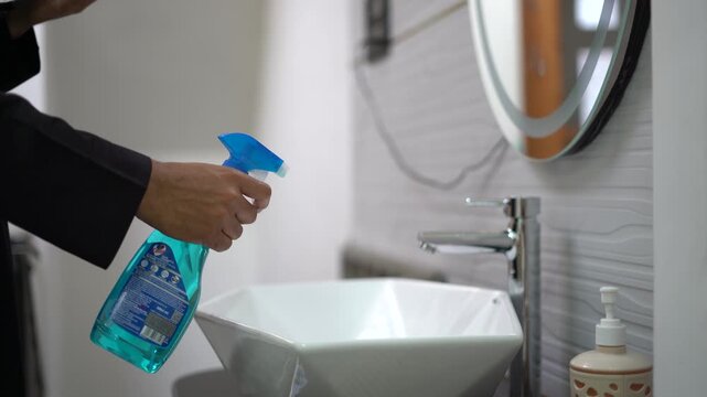 4K Close up of man in black jubbah using blue cleaning spray on modern bathroom sink, house cleaning and sanitization concept.