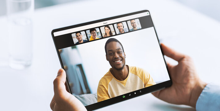 A young doctor is engaged in an online consultation. He holds a tablet with a video call on the screen featuring a smiling patient. The setting is a bright room with a white table.