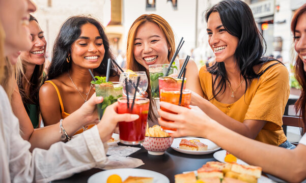 Happy people toasting cocktail glasses sitting at bar table - Cheerful friends enjoying happy hour at pub restaurant balcony - Food and beverage life style concept