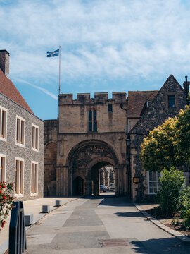 View of aged stone buildings flank a cobbled path leading under a medieval archway topped with a Scottish flag against a bright blue sky, Canterbury, England, United Kingdom.