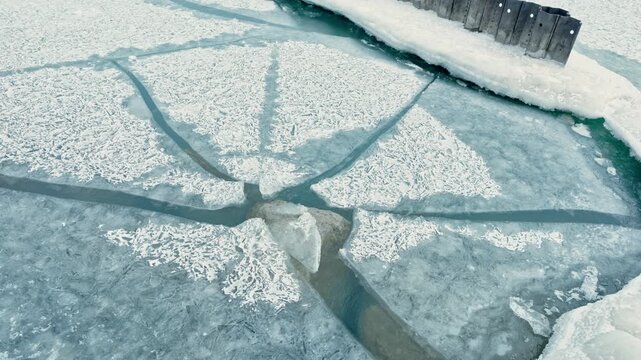 Frozen surface shows cracks and patterns with dark blue water visible underneath. The scene captures a cold winter day with a natural landscape surrounding the frozen area.