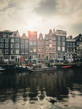 View of the canal reflecting the vibrant facades of historic buildings under a cloudy sky, punctuated by the bright sun, Amsterdam-centrum, Noord-Holland, Netherlands.