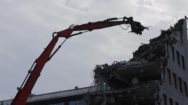 A giant demolition crane tears down an office building piece by piece, its powerful claw gripping and dismantling each section with force.