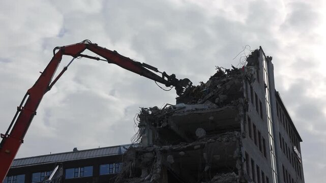 A giant demolition crane tears down an office building piece by piece, its powerful claw gripping and dismantling each section with force.