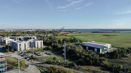 Fototapeta premium Industrial warehouses and apartments near Morrison Dr sit beside rail lines leading toward Cooper River waterfront. Sunlit rooftops, grassy fields, distant port structures in South Carolina