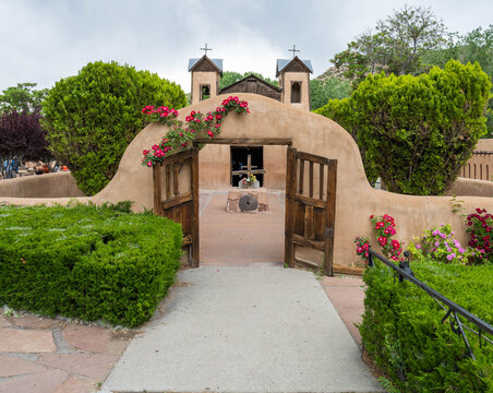 View of a historic adobe church framed by a weathered archway adorned with vibrant roses, casting shadows on the stone path, Santa Fe, New Mexico, United States.
