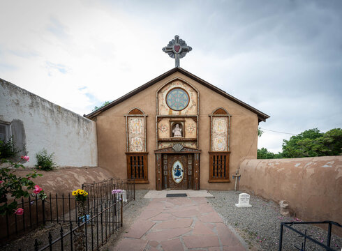 View of a quaint adobe church stands as a beacon of faith under a cloudy sky, its rustic charm accentuated by the surrounding walls, Santa Fe, United States.