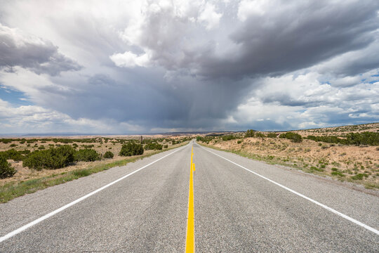 View of a long, gray asphalt road stretching into the horizon beneath a dramatic sky with gathering storm clouds, Santa Fe, New Mexico, United States.