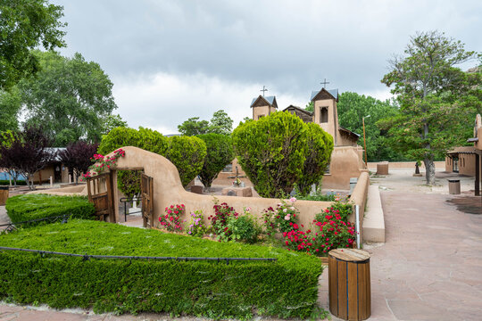 View of the historic adobe church surrounded by vibrant roses and lush greenery under a cloudy sky, embodying the serene beauty, Santa Fe, New Mexico, United States.