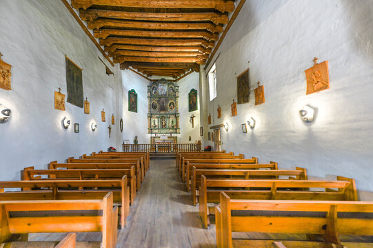 View of wooden pews aligned in a simple chapel under a beamed ceiling, walls adorned with religious art, casting a warm glow in Santa Fe, New Mexico, United States.