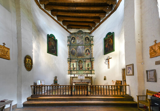 View of the historic chapel interior reveals rustic wooden beams and intricate carvings, casting shadows on the whitewashed walls, Santa Fe, New Mexico, United States.