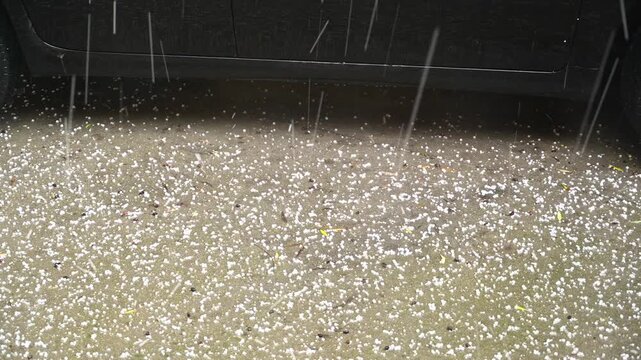 heavy hail falling on asphalt near a parked car. Hailstones bounce on the road surface creating a dramatic weather scene with impact, accumulation and intense atmospheric conditions.