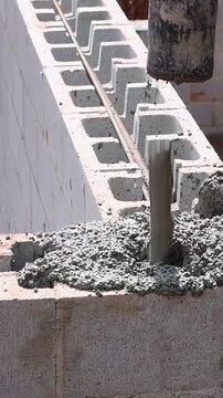 Worker stands on wall, directing concrete into block structure using pipe while surrounded by equipment.