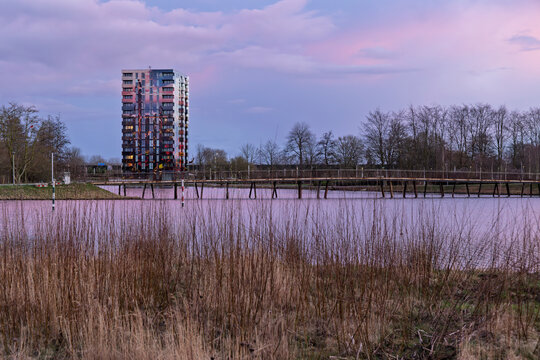 Almere, Netherlands. 14 March 2026. Modern Residential Tower Flores at Floriade Park, During Pink Sunset with Reflection on Weerwater Lake and Wooden Bridge