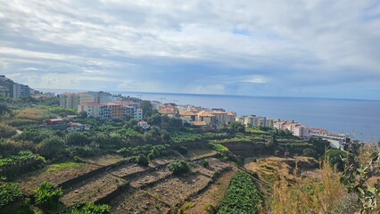 Panoramic view of Funchal, Madeira with terraced farmland, hillside homes and the Atlantic Ocean under a soft cloudy sky, showcasing the island’s coastal landscape. © Sebastiaan89