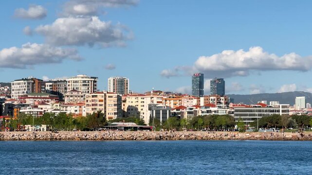 A sunny, clear day at Maltepe coast with fluffy white clouds in the sky. A serene coastal cityscape with modern buildings and people enjoying the seaside in Istanbul
