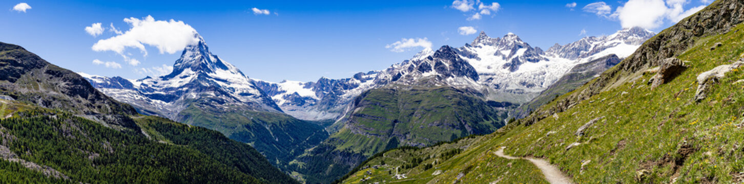 View of jagged, snow-capped peaks pierce the vibrant blue sky, contrasting against the lush green valleys and winding trails, Zermatt, Valais, Switzerland.
