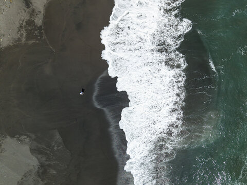 Aerial view of dark sands meet the turquoise waters, where frothy white waves crash, contrasting sharply with the dark beach, Pandansari Beach, Bantul Regency, Daerah Istimewa Yogyakarta, Indonesia.