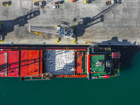 Aerial view of a cargo ship with a vibrant red deck loaded with white cargo, docked at the Ortona port with teal waters, Ortona, Abruzzo, Italy.