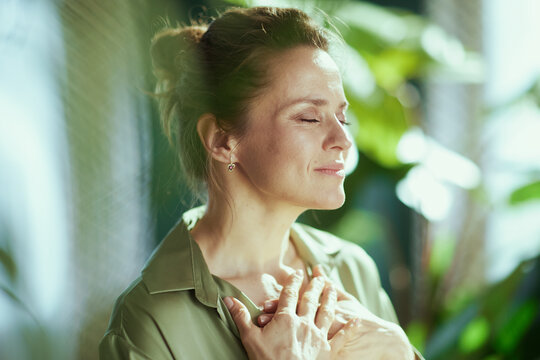 A woman stands peacefully with eyes closed and hands on her chest, finding a quiet moment of reflection and self-care in a lush, natural interior.