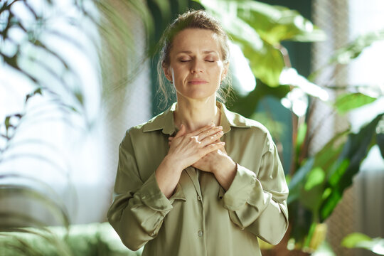 A middle-aged woman stands with her hands over her heart and eyes closed, engaging in a moment of self-compassion and emotional reset surrounded by greenery.