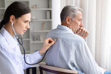 Fototapeta premium Female doctor examining senior patients lungs with stethoscope on back while patient coughs. Healthcare professional assessing respiratory condition during clinical checkup