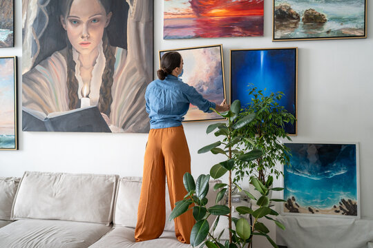 Successful woman painter arranging wall painting exhibition at studio loft apartment, stands on couch adjusting canvas on wall. Skillful female painter art lover displaying various artworks at home.