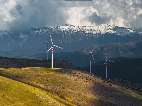 Aerial view of wind turbines standing tall against the backdrop of snow-capped mountains under a dramatic sky, Cocullo, Abruzzo, Italy.