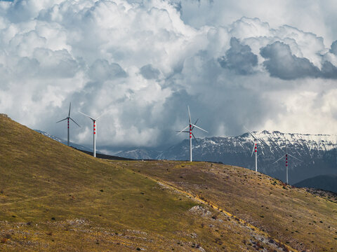 Aerial view of wind turbines standing tall against a backdrop of snow-dusted mountains and dramatic, cloud-filled skies, Cocullo, Abruzzo, Italy.