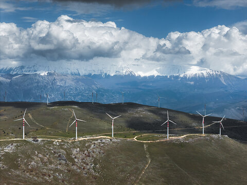 Aerial view of wind turbines standing tall against the backdrop of snow-capped mountains under a sky heavy with dramatic clouds, Cocullo, Abruzzo, Italy.