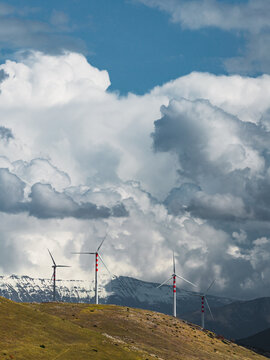 Aerial view of wind turbines standing tall against a backdrop of dramatic, swirling clouds and distant snow-capped mountains, Cocullo, Abruzzo, Italy.