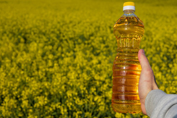 A man stands in a rapeseed field holding a clear bottle filled with rapeseed oil under the sun. The field is covered in yellow flowers