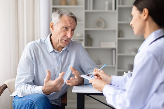 Senior male patient explaining symptoms to female doctor during clinic consultation. Healthcare professional taking notes and assessing medical condition