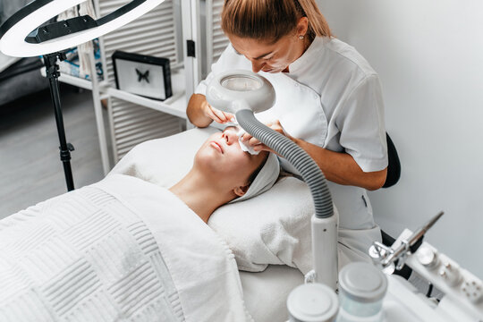 Female cosmetologist using magnifying lamp to perform skin cleaning and facial treatment on woman in professional beauty clinic.