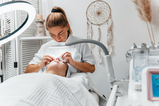 Female cosmetologist using magnifying lamp to perform skin cleaning and facial treatment on woman in professional beauty clinic.