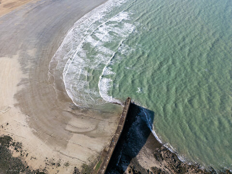 Aerial view of a stone pier reaching into the teal ocean where waves crash against the sandy shore, Pleneuf-Val-Andre, Brittany, France.