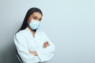 Woman in medical coat with mask stands with arms crossed, looking directly at the camera in a neutral setting during daytime © verona_studio