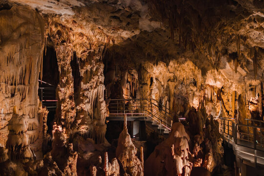 Wide view of Petralona Cave in Halkidiki, Greece, showing modern metal walkways and staircases winding among massive stalactite and stalagmite formations under warm lighting.