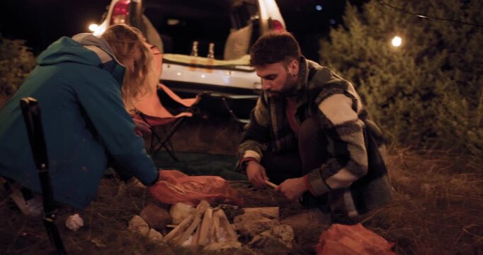 Couple preparing food under string lights by car in dark outdoor area at night near trees
