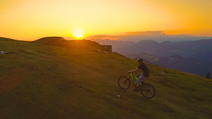 Fototapeta premium DRONE: Golden sunset illuminates the way for mountain biker pedalling uphill.