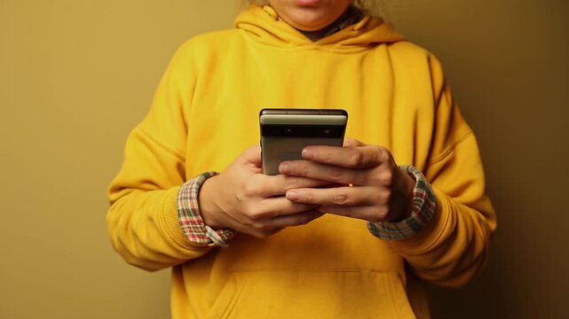 Close-up of a person in a yellow hoodie holding and using a smartphone. Person texting, browsing social media, or using a mobile app on a plain background. Digital lifestyle and communication concept