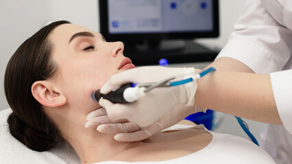 Female patient receiving hydrafacial treatment in a beauty clinic, technician using a handheld device on her cheek, computer monitor visible in the background © wedmoments.stock