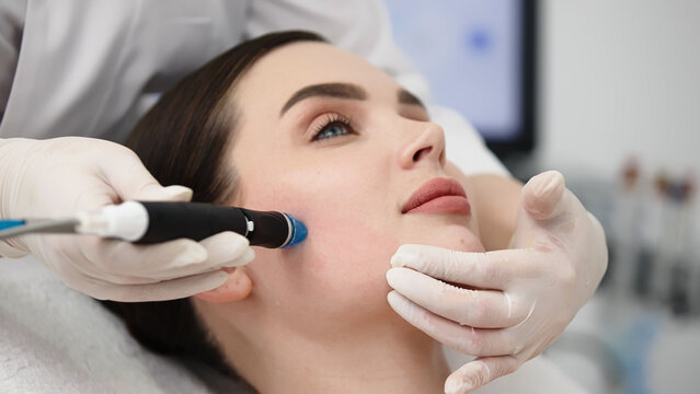 Female patient receiving hydrafacial treatment in a beauty clinic, with a technician applying a device to her face while wearing gloves and a white coat