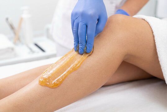 Beautician in blue gloves applying sweet sugaring paste onto a female leg for hair removal