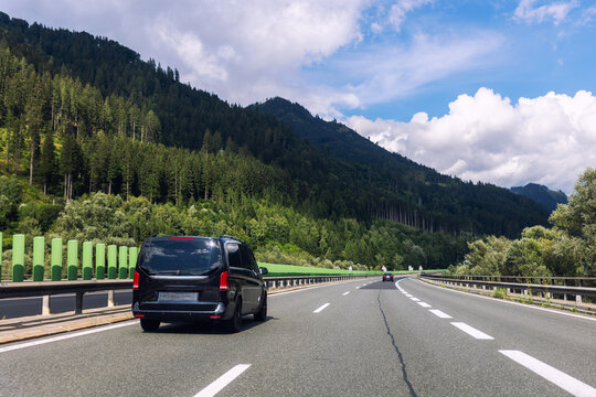 Luxury black minivan driving along alpine highway between green mountains blue sky. Comfort vip van transfer transportation passenger service, Airport shuttle taxi European mountain resorts