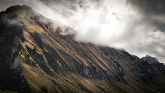 Aerial view of dramatic sunlight illuminating the rugged, shadowed face of Pic de Jallouvre, shrouded in ethereal clouds, Glieres-Val-de-Borne, Auvergne-Rhone-Alpes, France.