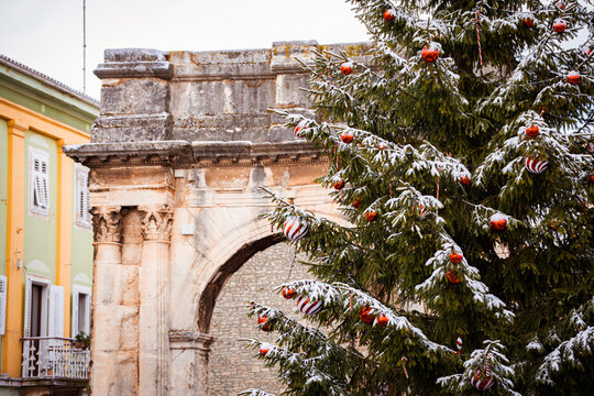 View of snow-dusted Christmas tree ornaments sparkle against ancient stone archways, a vibrant contrast of festive spirit and timeless architecture, Pula, Croatia.